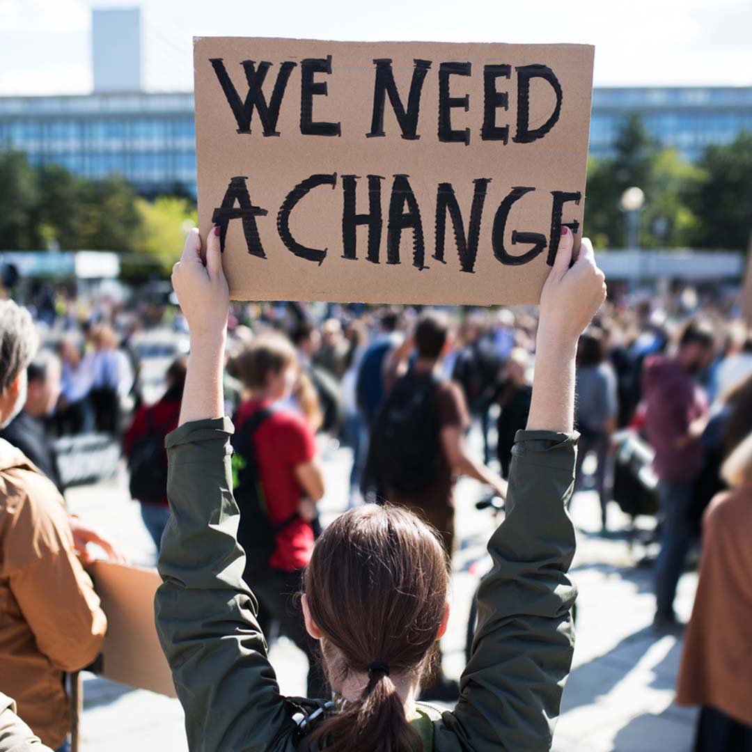 A person holding a protest sign reading "We Ned A Change"