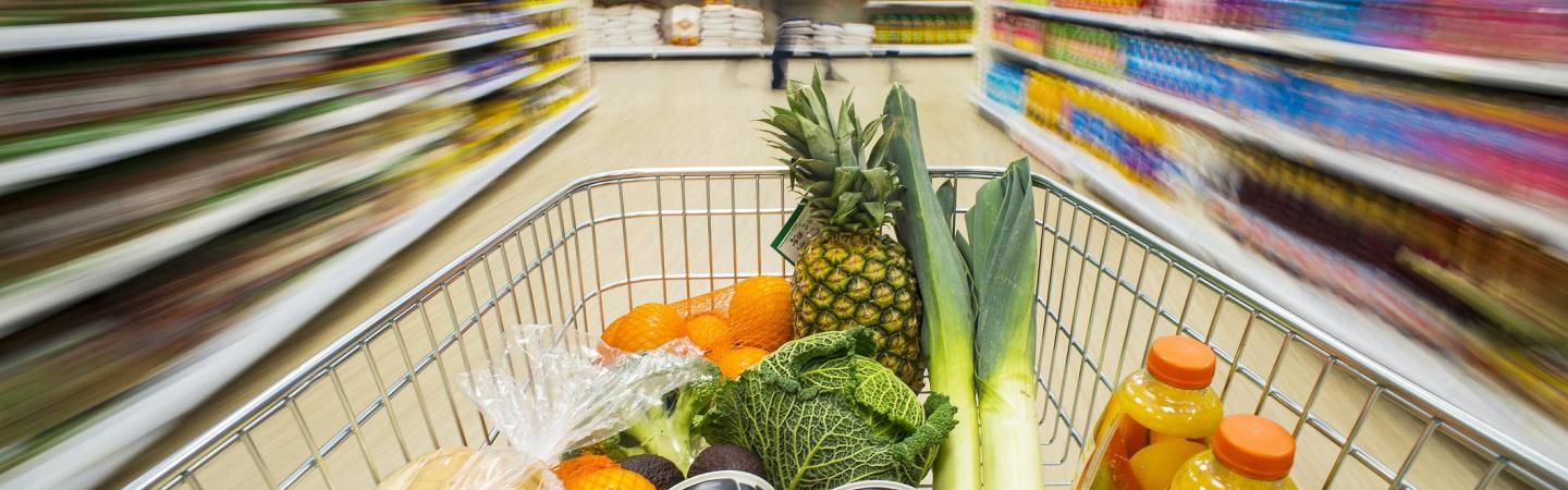 A grocery cart filled with produce and canned foods moving down a grocery store aisle