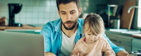 father sitting at table with toddler on lap next to laptop, paperwork, and calcuator