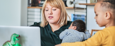 image of mother holding infant while working on laptop while toddler looks on