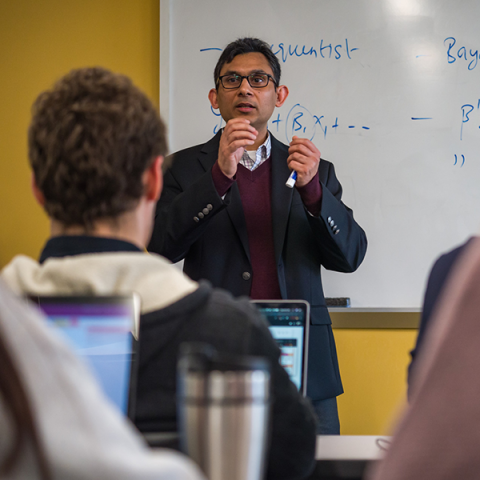 Photo of an instructor teaching a class while standing in front of a white bard with symbols written on it.