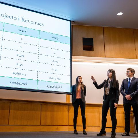 Three students standing in front of a screen with revenue information projected upon it