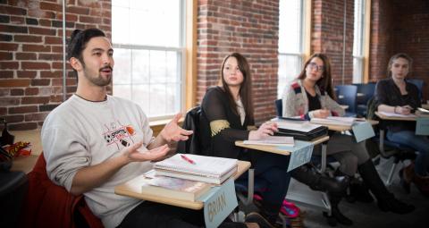 A male student sitting in a class speaking while female students look on.